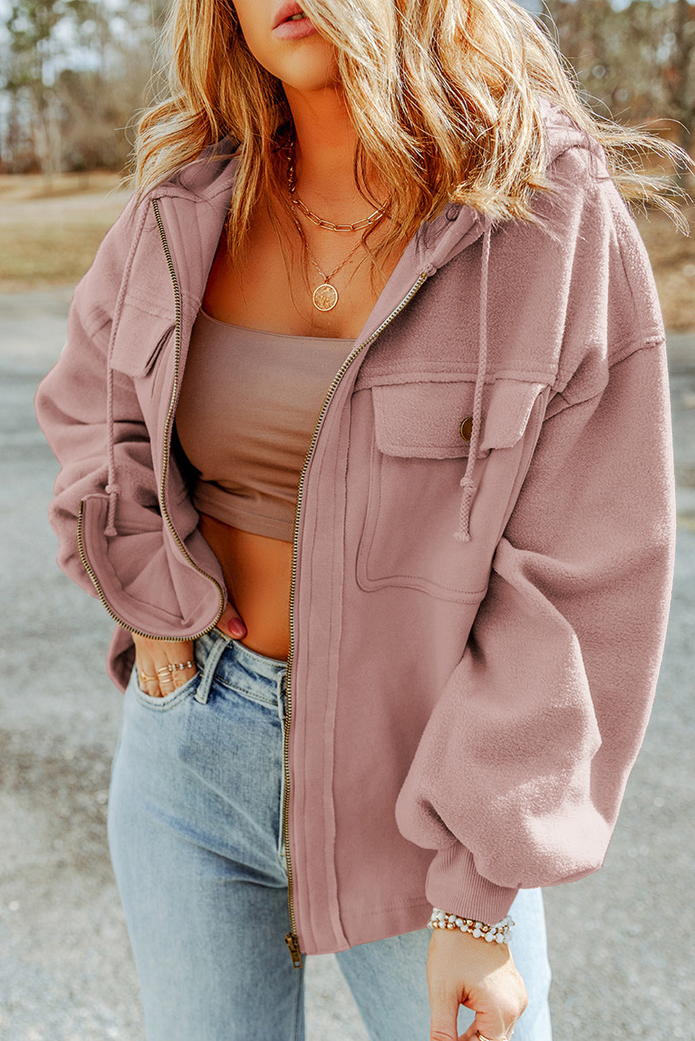 Woman wearing a pink jacket, brown tank top, and light blue jeans, accessorized with gold necklaces and bracelets, set against an outdoor backdrop.