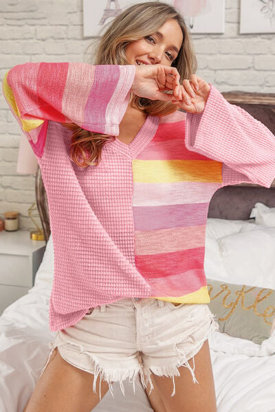 A woman in a playful pose wearing a pink, yellow, and red striped sweater and white frayed denim shorts, in a cozy bedroom with a hint of decor in the background.