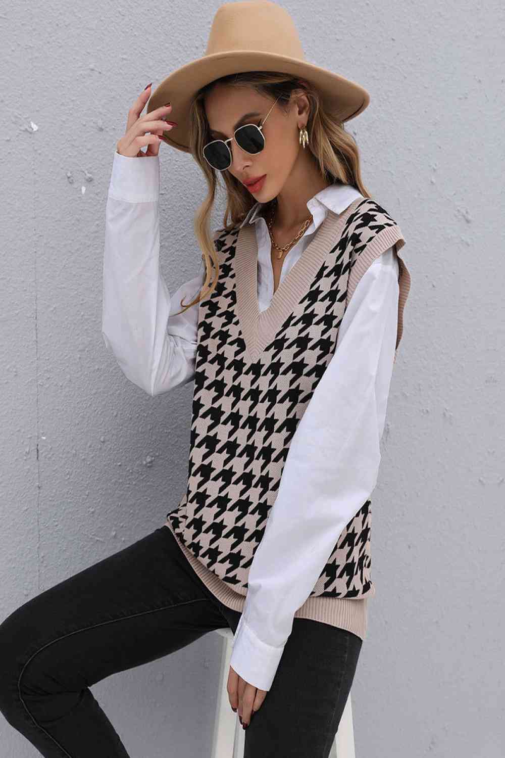 Woman wearing a beige houndstooth sweater vest over a white button-down shirt, paired with black jeans and accessorized with a beige hat, sunglasses, and gold jewelry.