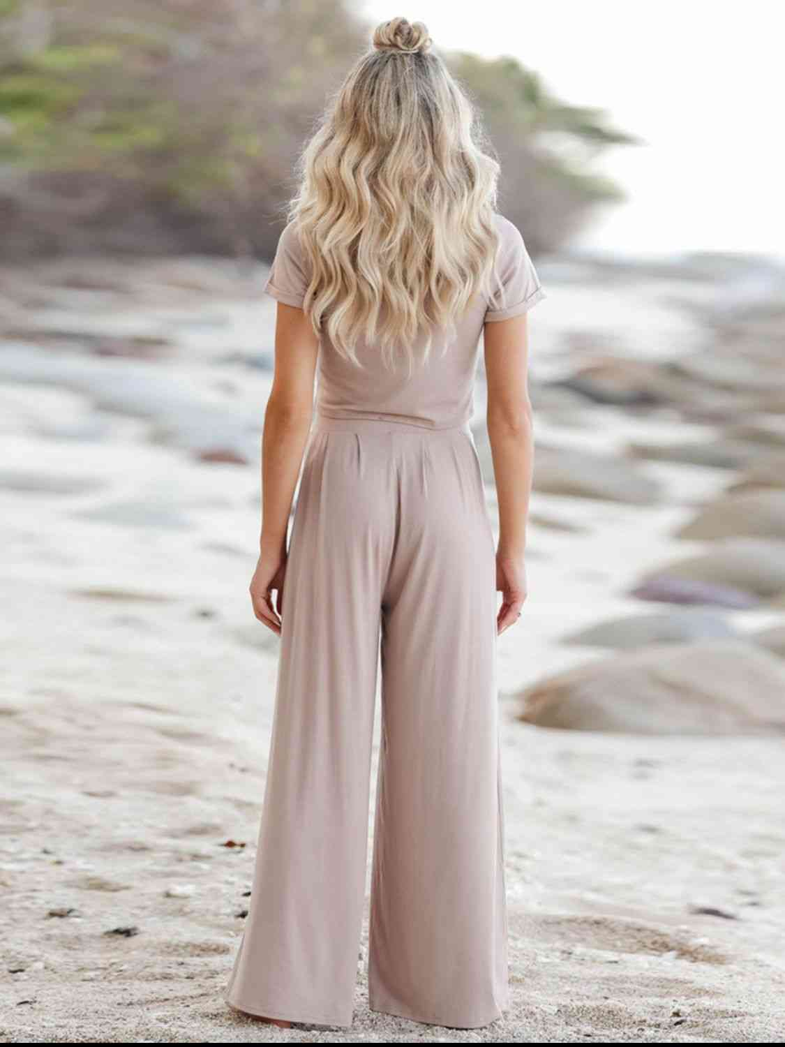 Woman standing on a sandy beach, seen from behind, wearing a tan shirt and pants with her hair down, looking forward.