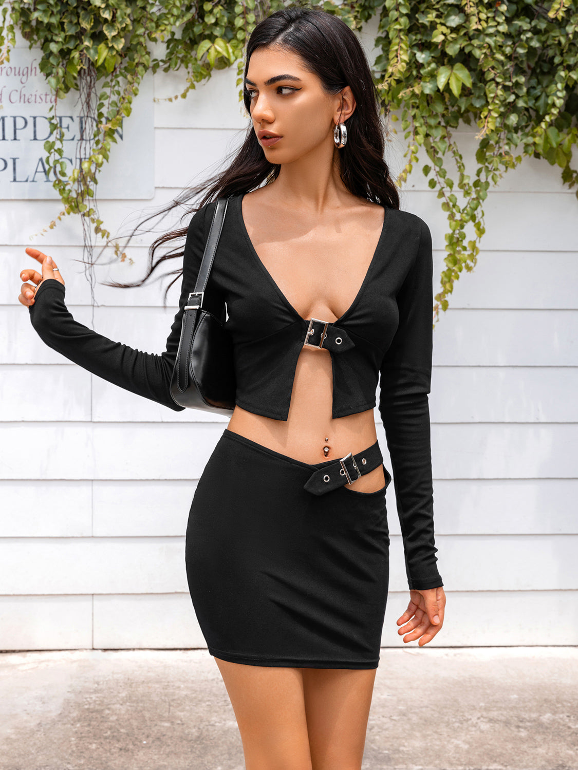 A side view of a model in a black two-piece set with a long-sleeve cropped top and a mini skirt featuring cutout and buckle details, showcasing the outfit's flattering fit.