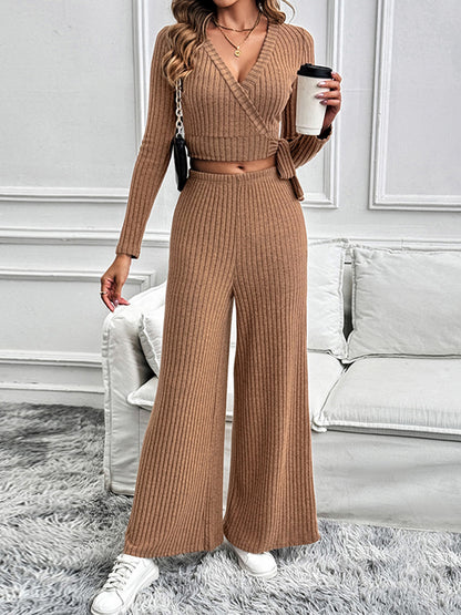 A model stands in a stylish ribbed knit two-piece lounge set in camel, holding a coffee cup. The outfit features a wrap-style surplice top with a tied waist and wide-leg pants, paired with white sneakers.