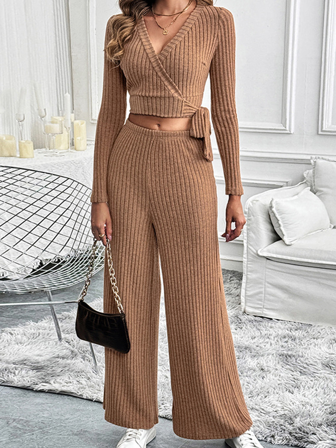 Front view of a model wearing a camel-colored ribbed knit two-piece lounge set with a wrap-style top and wide-leg pants, accessorized with a black purse and white sneakers.