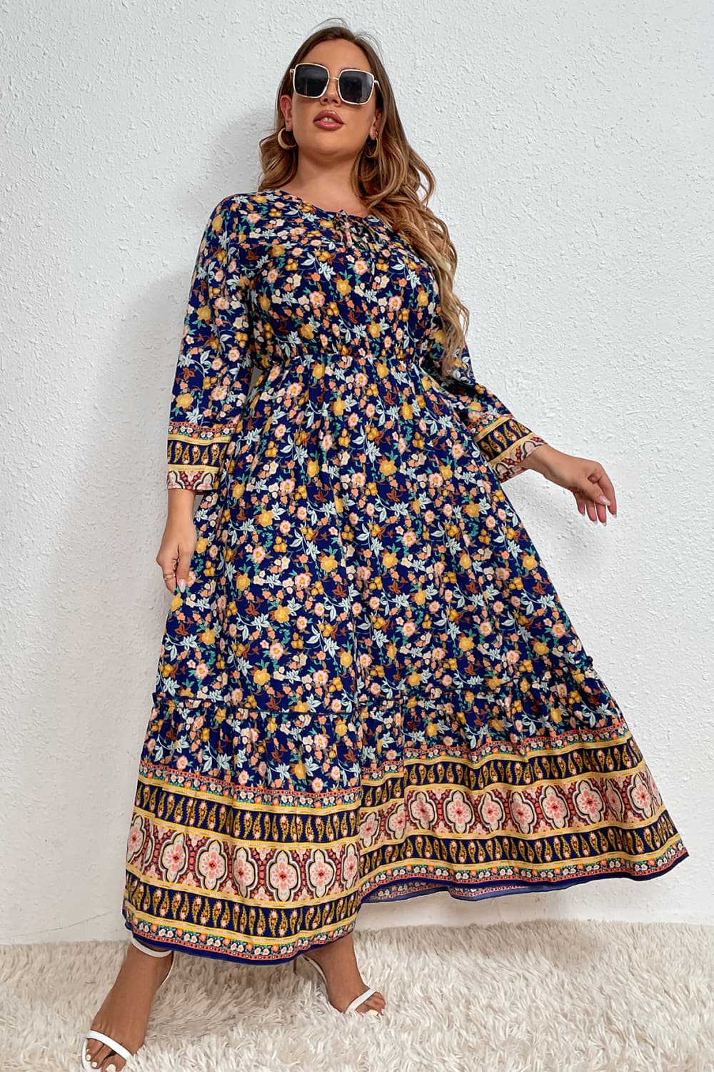 Stylish woman in a floral bohemian dress with intricate border patterns, complemented by sunglasses, set against a white wall and standing on a plush white rug.