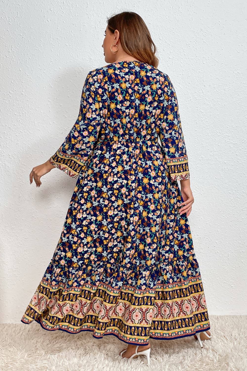 Stylish woman in a floral bohemian dress with intricate border patterns, complemented by sunglasses, set against a white wall and standing on a plush white rug.