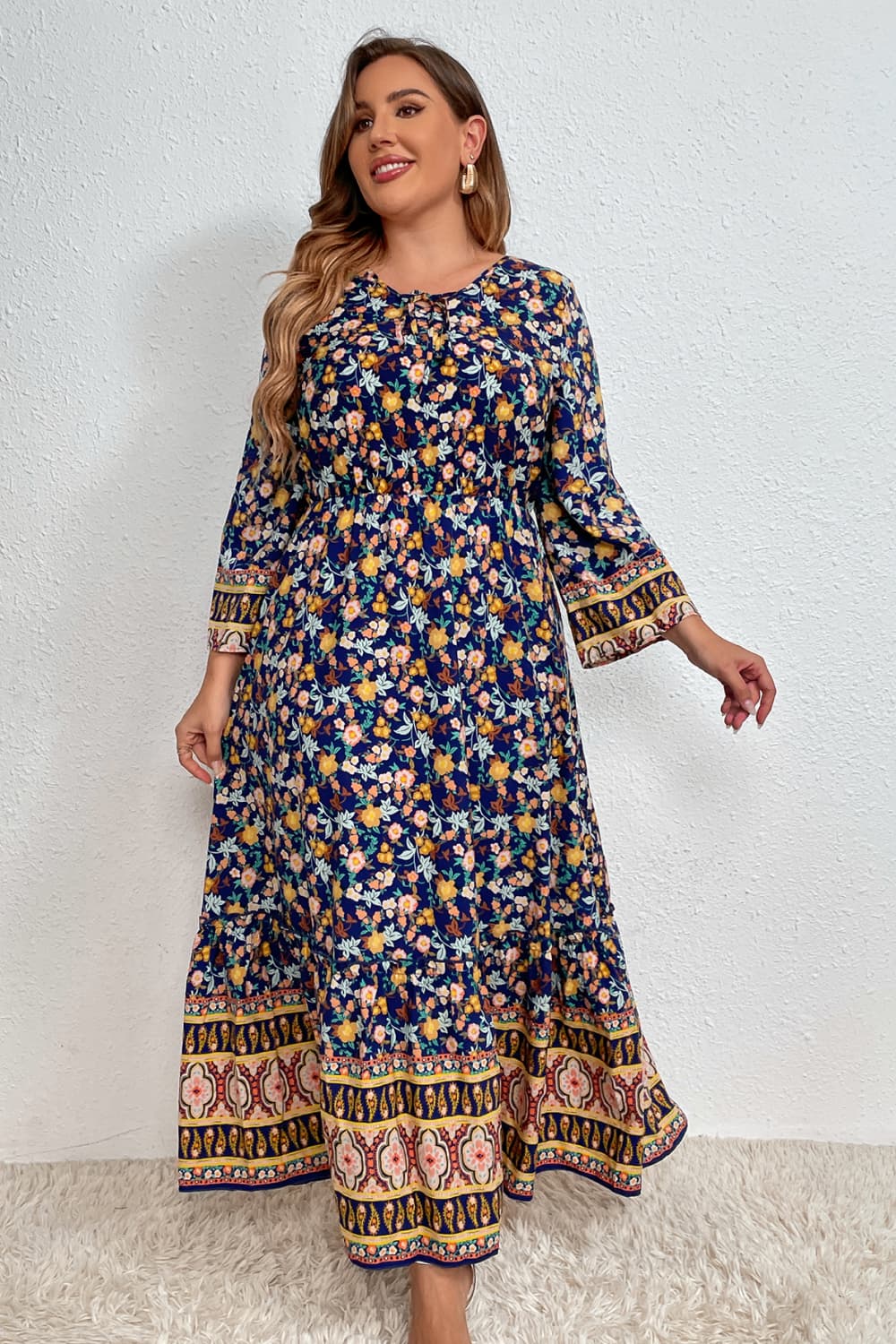 Stylish woman in a floral bohemian dress with intricate border patterns, complemented by sunglasses, set against a white wall and standing on a plush white rug.