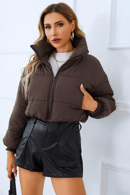  Woman in a brown puffer jacket, white sweater, and black leather shorts, paired with gold jewelry, against a white backdrop.