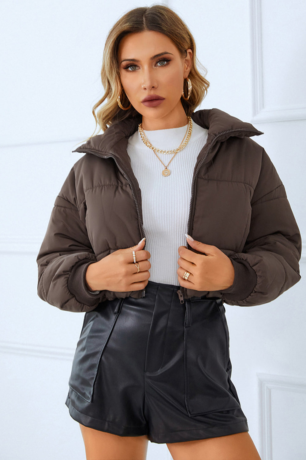 Woman in a brown puffer jacket, white sweater, and black leather shorts, paired with gold jewelry, against a white backdrop.