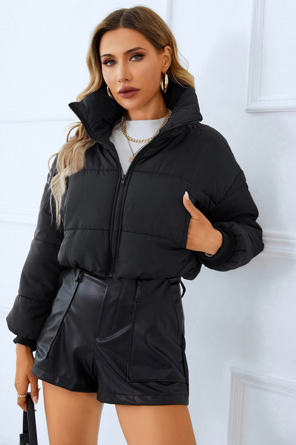 Woman in a black puffer jacket, white sweater, and black leather shorts, paired with gold jewelry, against a white backdrop.