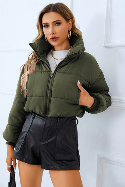 Woman in a green puffer jacket, white sweater, and black leather shorts, paired with gold jewelry, against a white backdrop.