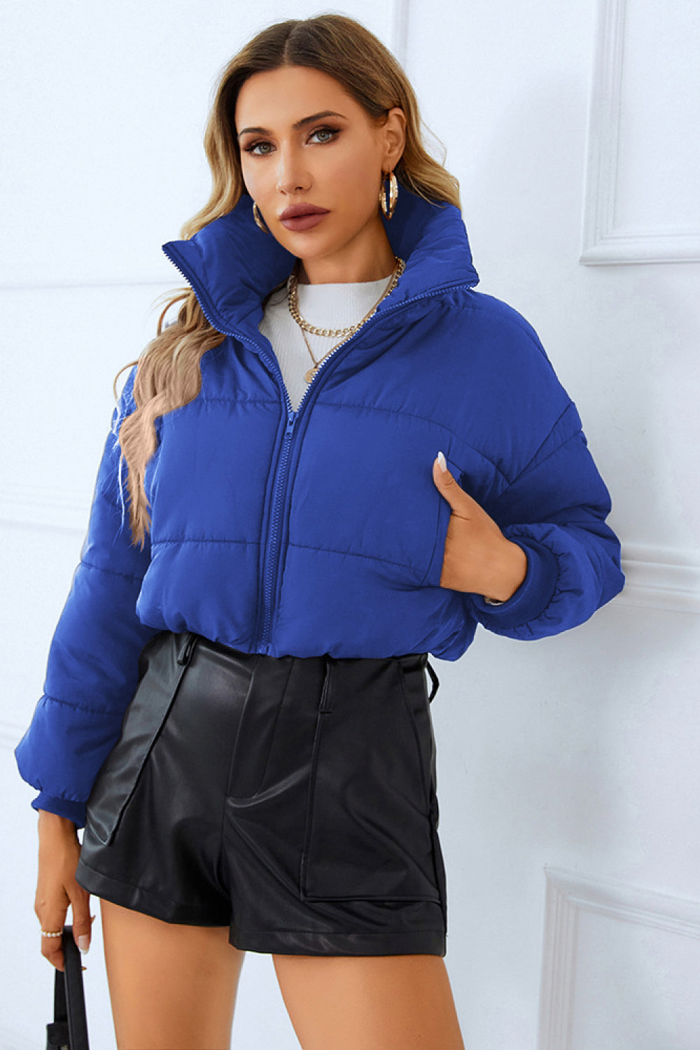 Woman in a blue puffer jacket, white sweater, and black leather shorts, paired with gold jewelry, against a white backdrop.