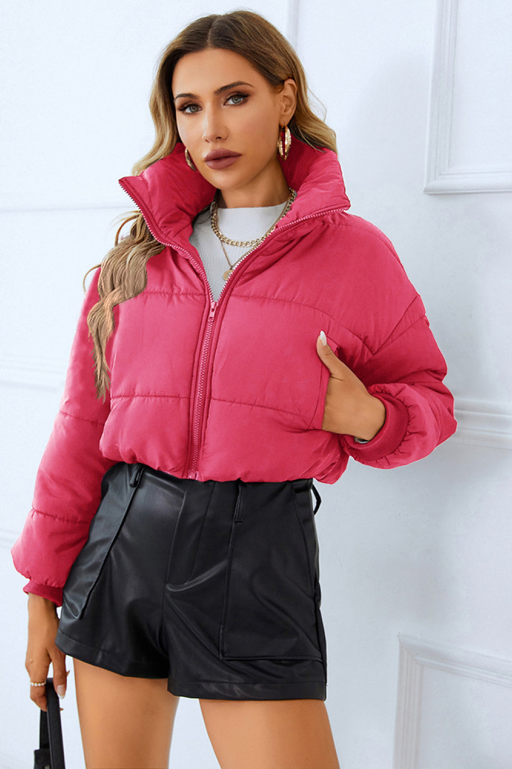 Woman in a dark pink puffer jacket, white sweater, and black leather shorts, paired with gold jewelry, against a white backdrop.