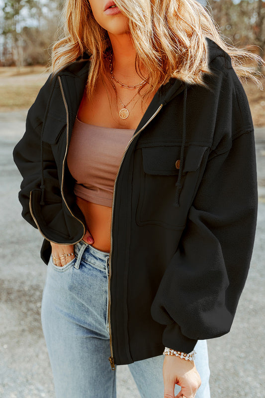 Woman wearing a black jacket, brown tank top, and light blue jeans, accessorized with gold necklaces and bracelets, set against an outdoor backdrop.