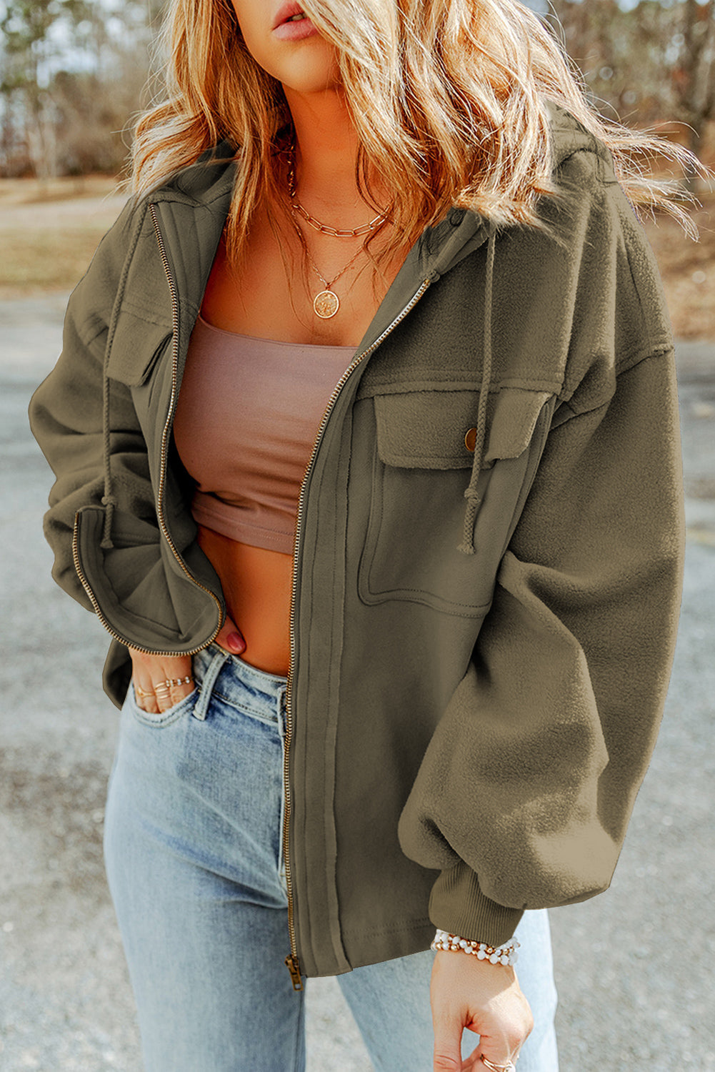 Woman wearing a green jacket, brown tank top, and light blue jeans, accessorized with gold necklaces and bracelets, set against an outdoor backdrop.