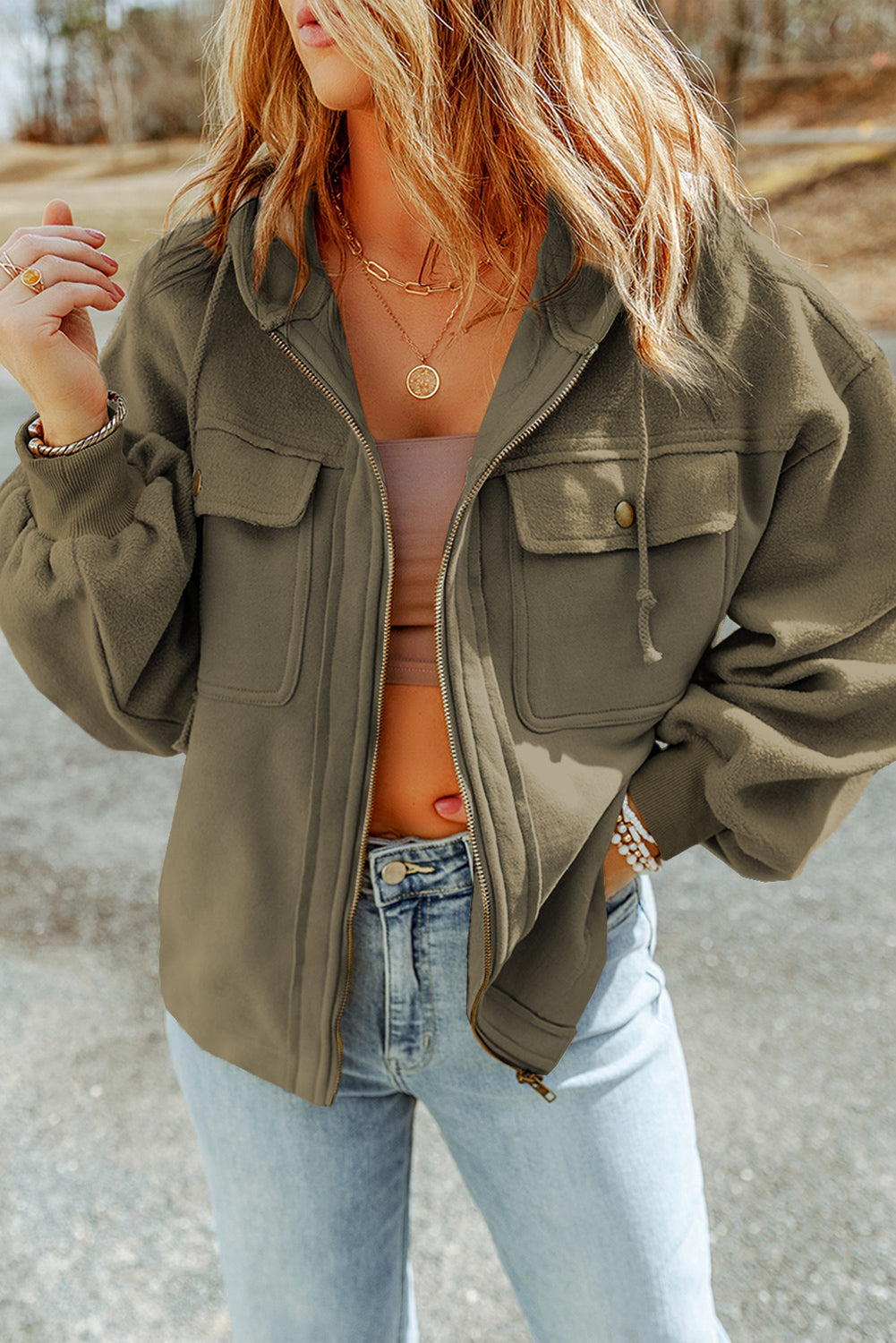 Woman wearing a green jacket, brown tank top, and light blue jeans, accessorized with gold necklaces and bracelets, set against an outdoor backdrop.