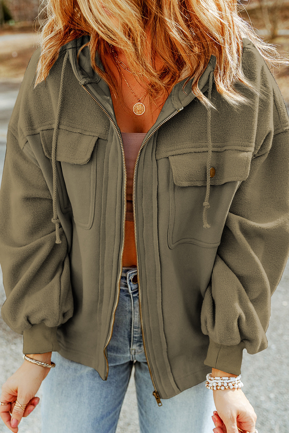 Woman wearing a green jacket, brown tank top, and light blue jeans, accessorized with gold necklaces and bracelets, set against an outdoor backdrop.