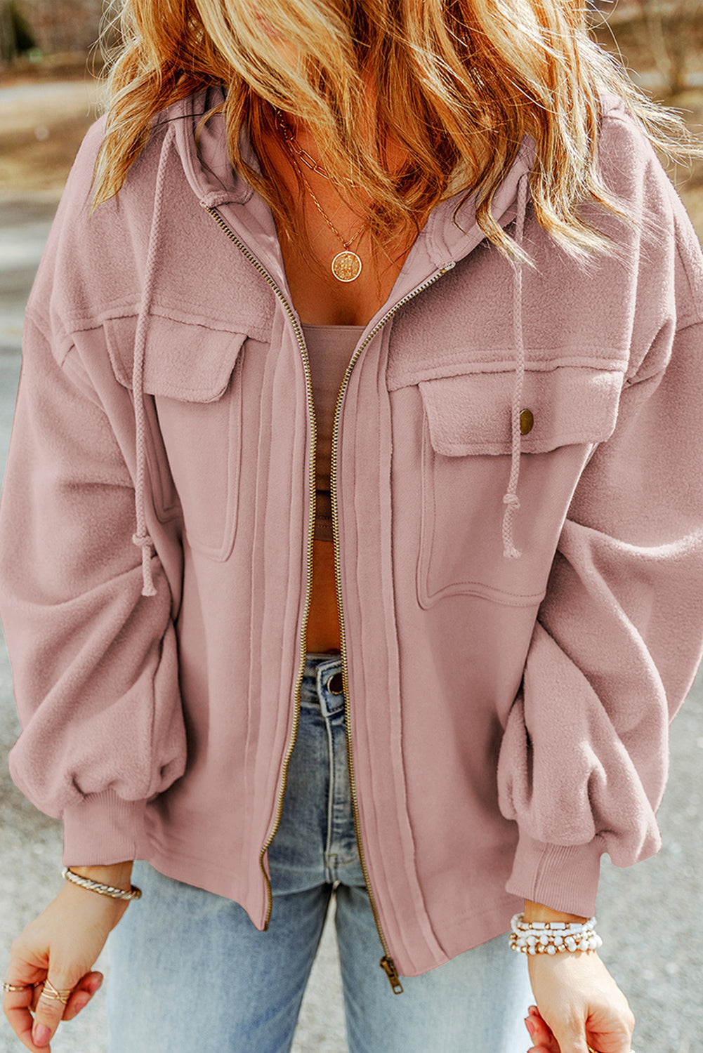 Woman wearing a pink jacket, brown tank top, and light blue jeans, accessorized with gold necklaces and bracelets, set against an outdoor backdrop.