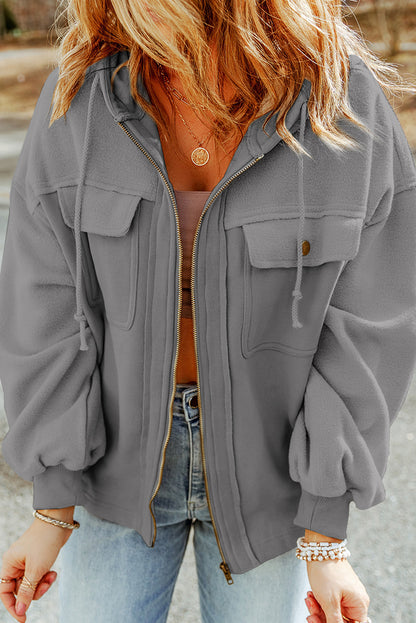 Woman wearing a gray jacket, brown tank top, and light blue jeans, accessorized with gold necklaces and bracelets, set against an outdoor backdrop.