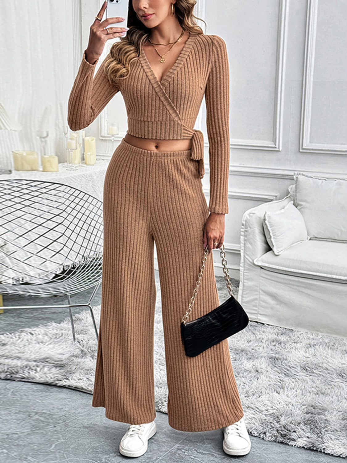 Front view of a model wearing a camel-colored ribbed knit two-piece lounge set with a wrap-style top and wide-leg pants, accessorized with a black purse and white sneakers.