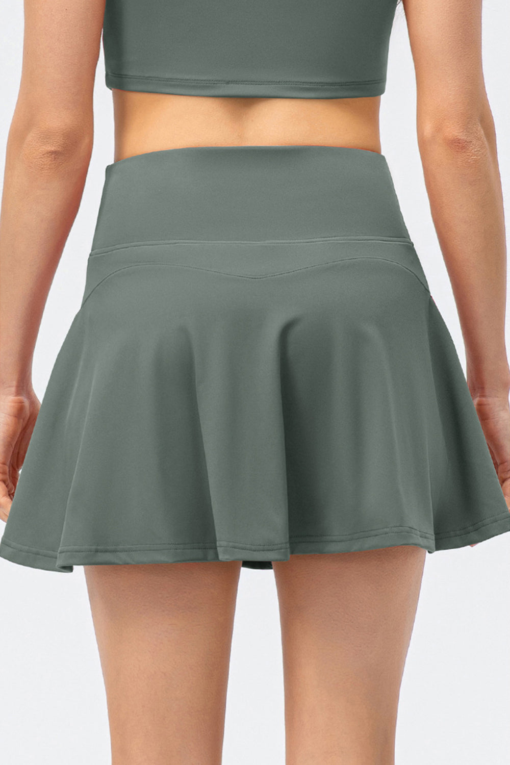 The back view of a model wearing a high-waist active skirt in muted green, showcasing the wide waistband and flared silhouette.