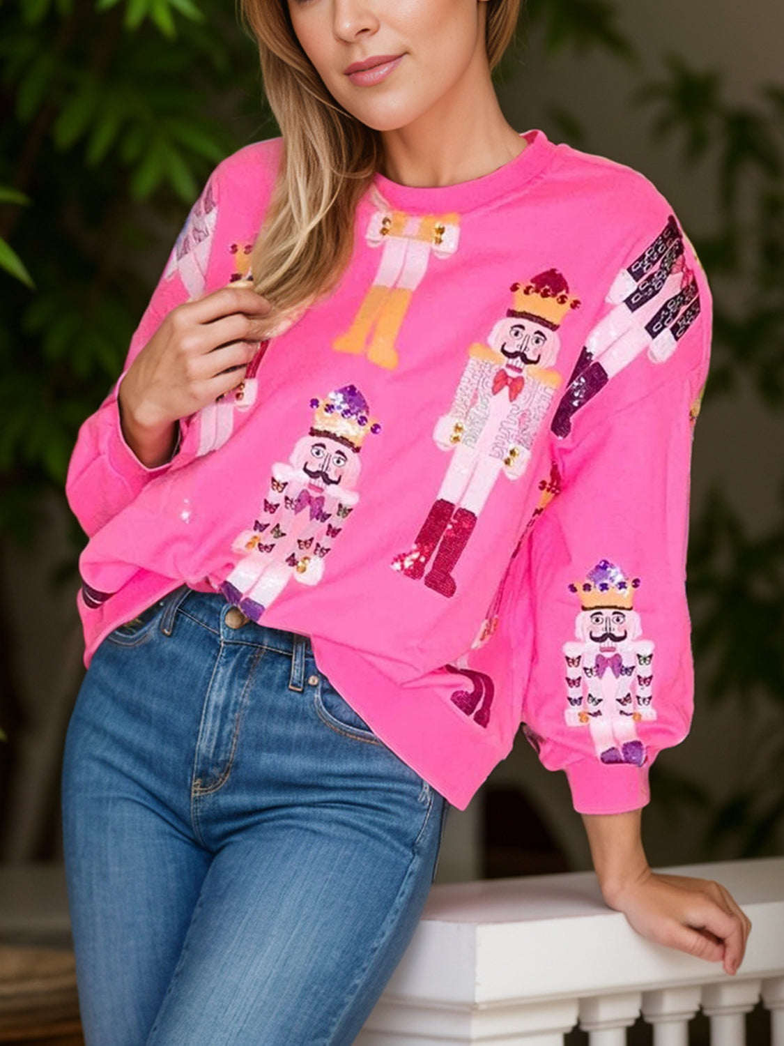 Woman wearing a bright pink sweatshirt with nutcracker designs, paired with blue jeans, posing indoors.