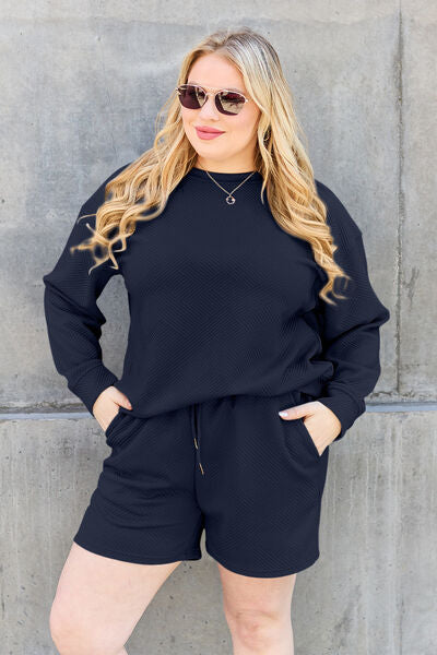 A woman in a casual dark blue loungewear set, comprising a sweatshirt and shorts, complemented with sunglasses and a necklace, posing confidently against a grey concrete wall.