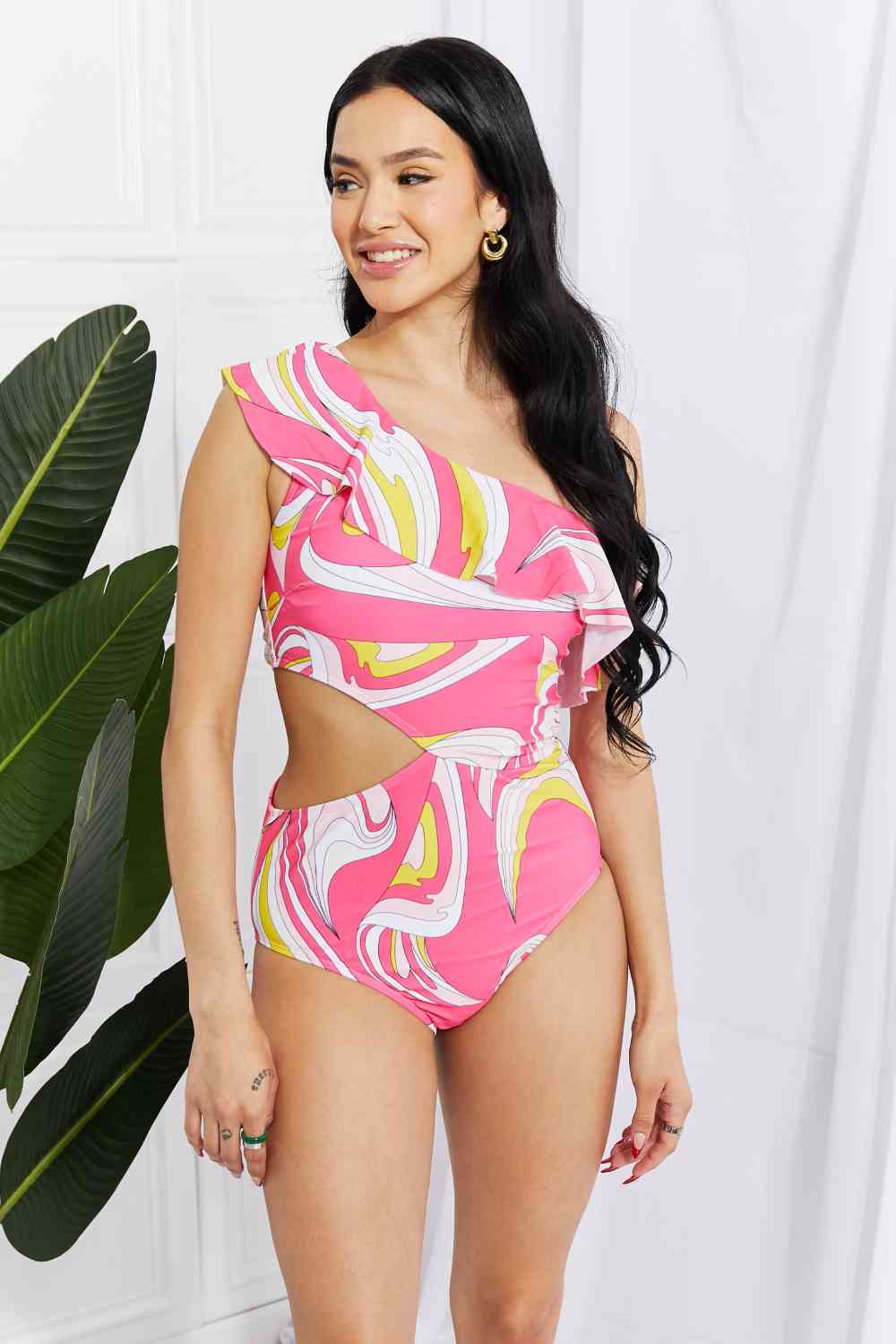 Woman wearing a bold pink one-shoulder cutout swimsuit with abstract print and ruffle detail, smiling and posing with hands on hips.