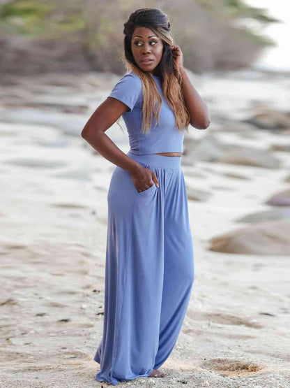 A woman looking away on a beach, wearing a blue cropped top and wide-leg pants, with one hand on her hip and the other touching her hair.