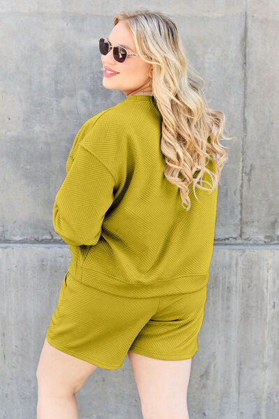 A woman viewed from behind, looking over her shoulder, wearing a yellow-green sweatshirt and shorts set, with her curly hair flowing, and wearing round sunglasses against a grey wall.