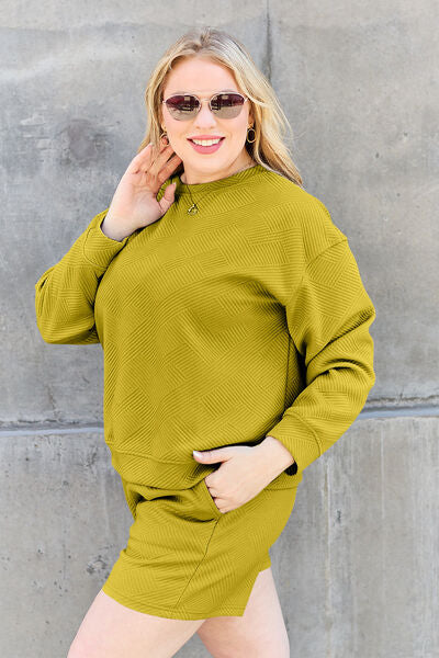 A woman with a smile, wearing a yellow-green textured loungewear set and sunglasses, touches her hair while posing against a textured concrete wall.