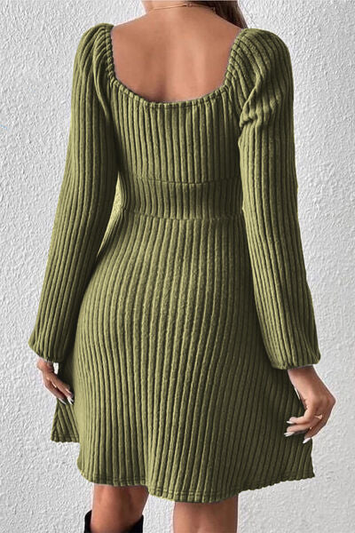 Woman facing away, wearing a ribbed dark green long-sleeve dress with a scooped back, standing against a white textured wall.