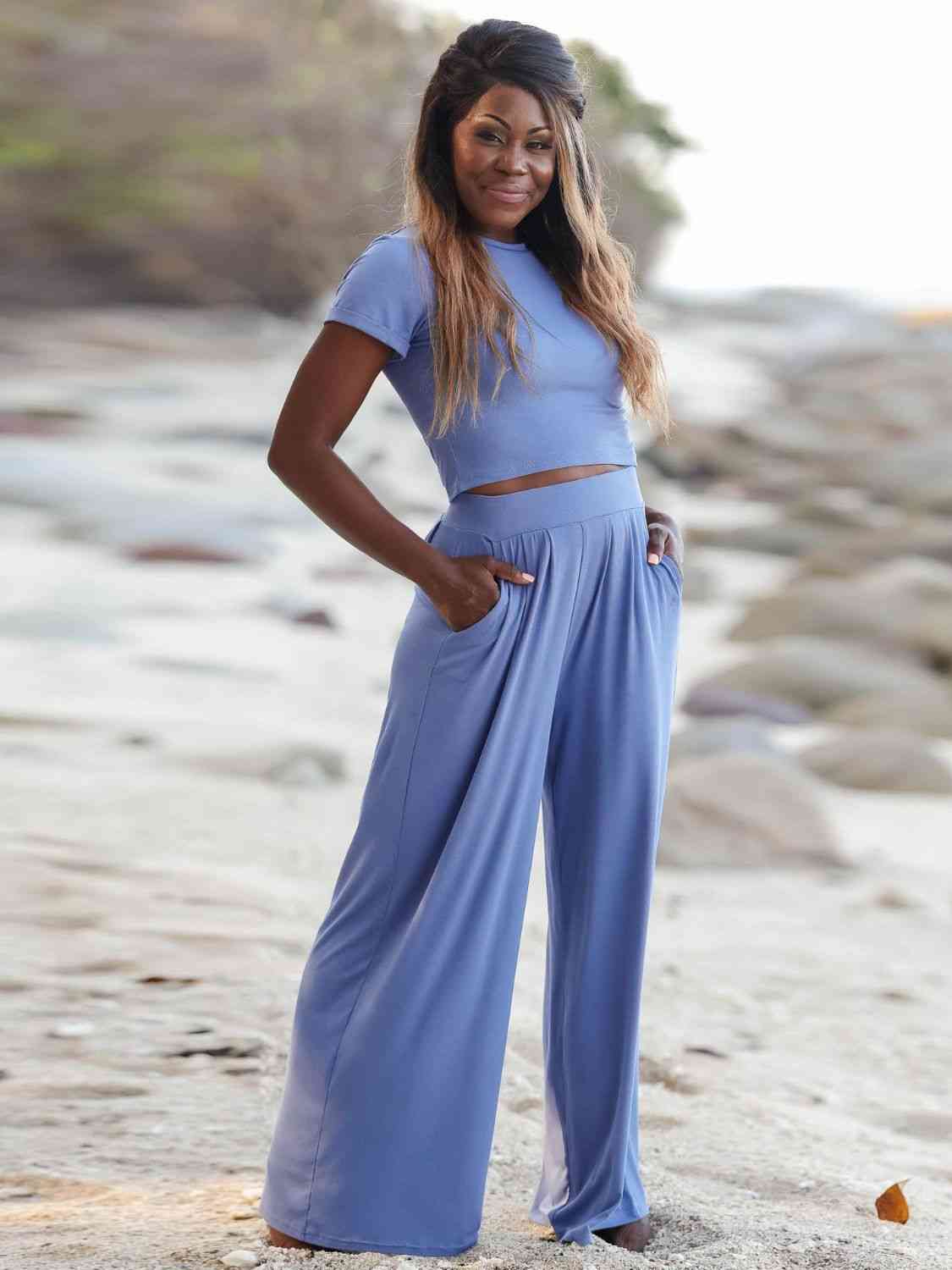 A woman smiling on a beach, wearing a blue cropped top and wide-leg pants, with her hand on her hip.