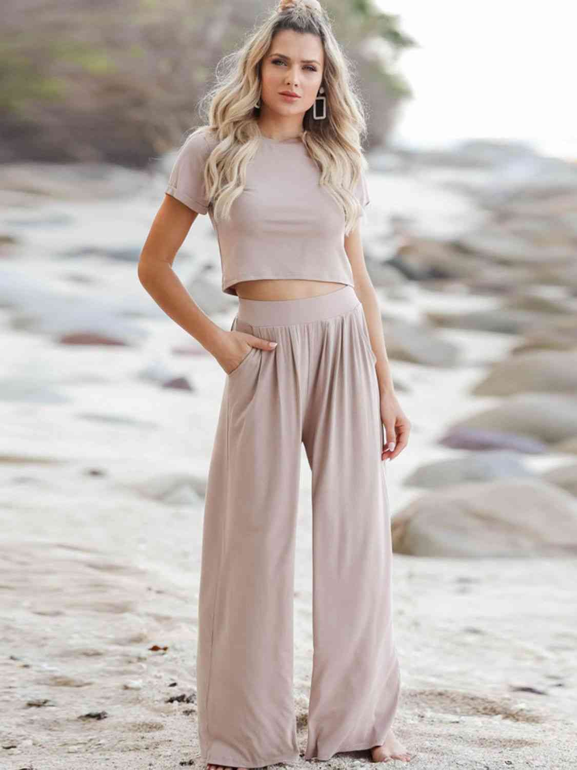 Woman on a sandy beach wearing a beige crop top and flowing high-waisted pants, looking forward with a thoughtful expression.