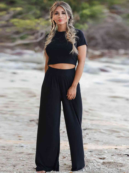 A woman smiling on a beach, wearing a black cropped top and wide-leg pants, with her hand on her hip.