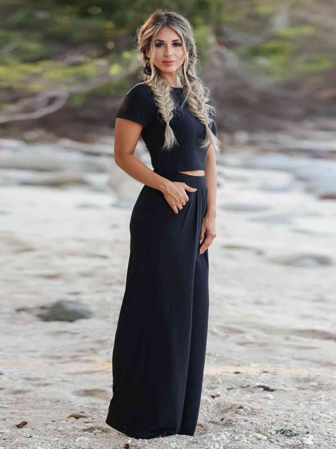 A woman smiling on a beach, wearing a black cropped top and wide-leg pants, with her hand on her hip.