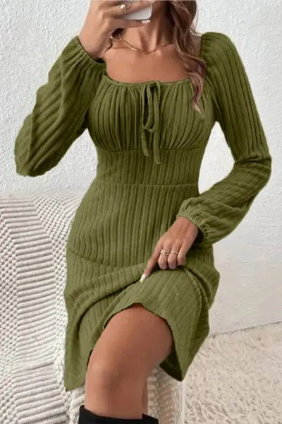 Woman in a moss green ribbed dress with a square neckline and drawstring detail, paired with a necklace, sitting and holding a cell phone. 