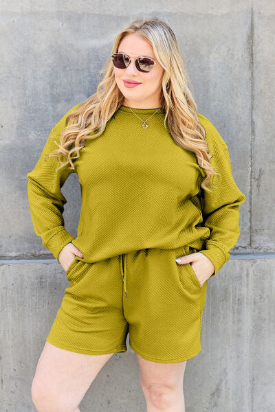 A woman in a casual yellow-green loungewear set, comprising a sweatshirt and shorts, complemented with sunglasses and a necklace, posing confidently against a grey concrete wall.