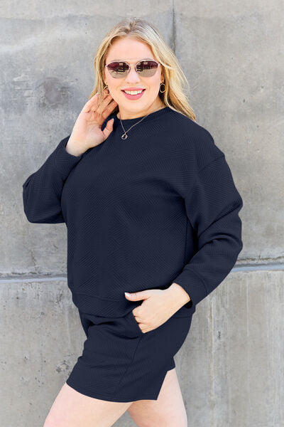 A woman with a smile, wearing a dark blue textured loungewear set and sunglasses, touches her hair while posing against a textured concrete wall.