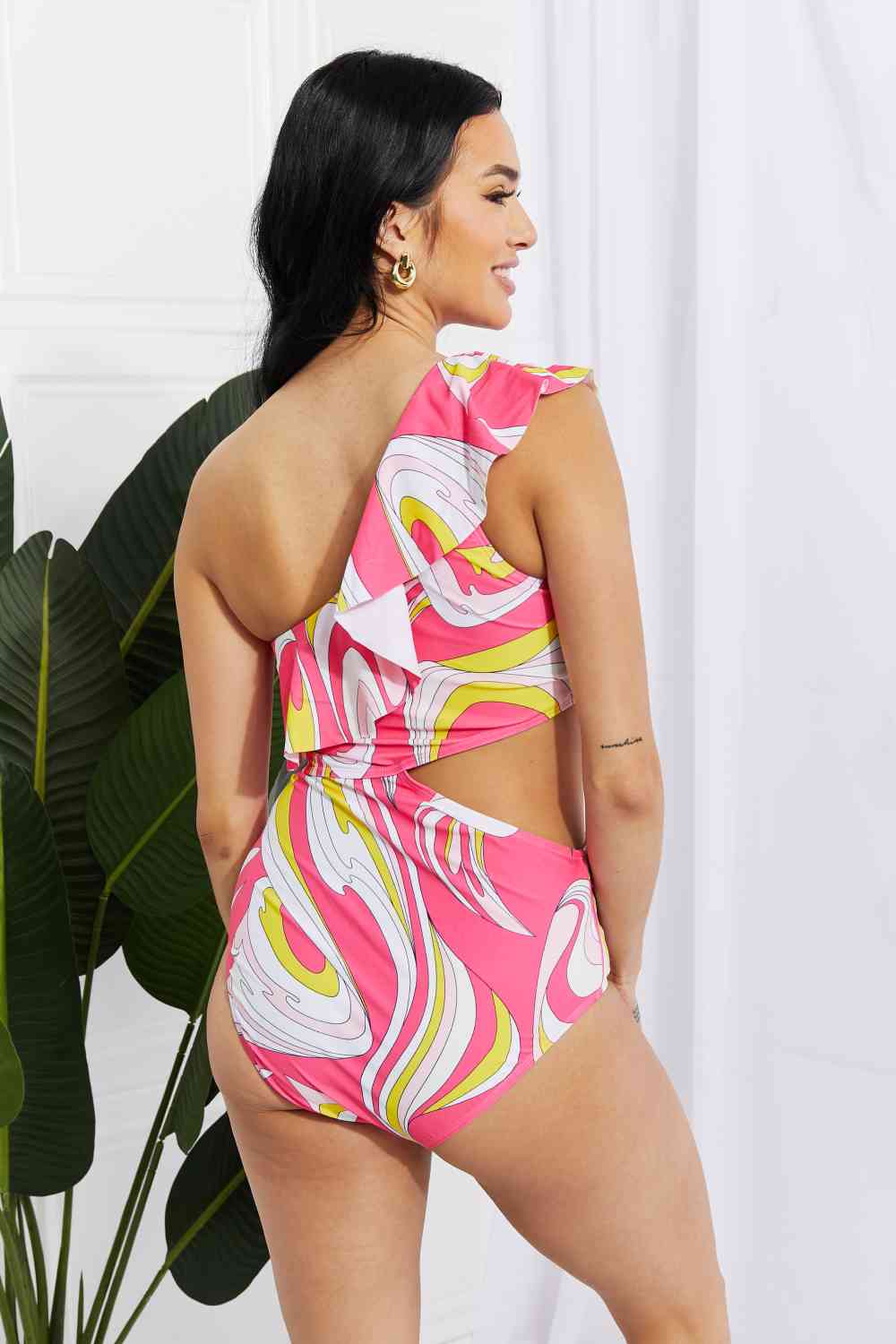 Woman showcasing the back of the pink asymmetric swimsuit, revealing the cutout design and ruffle detailing on the shoulder, standing near plants.